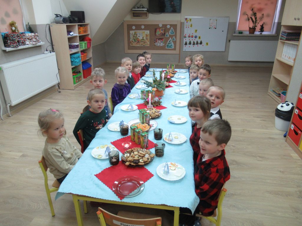 Dzieci z grupy 3-4 latków siedzą przy stołach nakrytych obrusami . Znajdują się na nich wigilijne smakołyki.