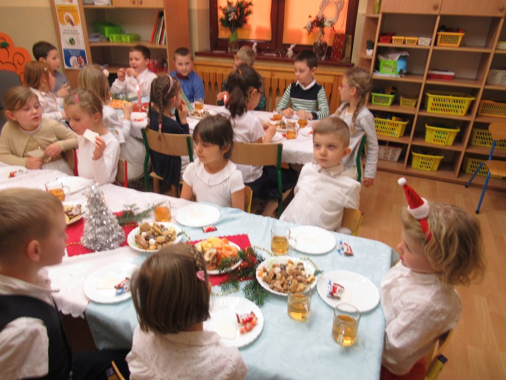 Wigilijka w grupie dzieci 6 letnich. Dzieci siedzą przy stołach ułożonych w podkowę, nakrytych obrusami i stroikami. Dziela sie opłatkiem i zajadają słodkie ciasteczka.