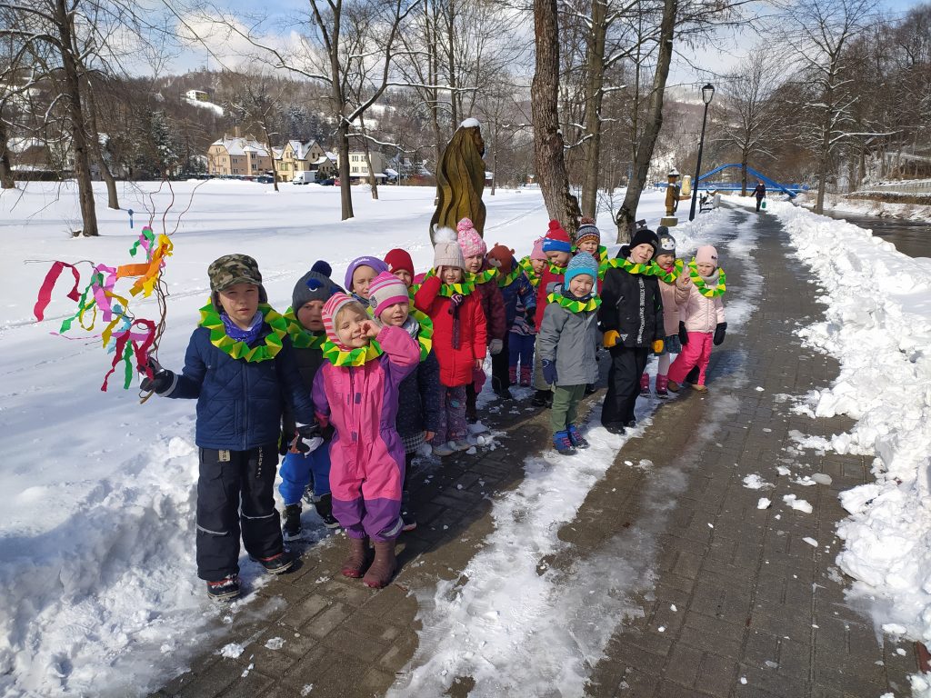 Zdjęcie grupy 3-4 latków ze spaceru z gaikiem. Dzieci idą w parach na Bulwarze. Pierwsza para trzyma  gaik z gałązki ozdobiony kolorowymi wstążkami z bibułki. Dzieci idą witać wiosnę. Po obu stronach Bulwaru śnieg.