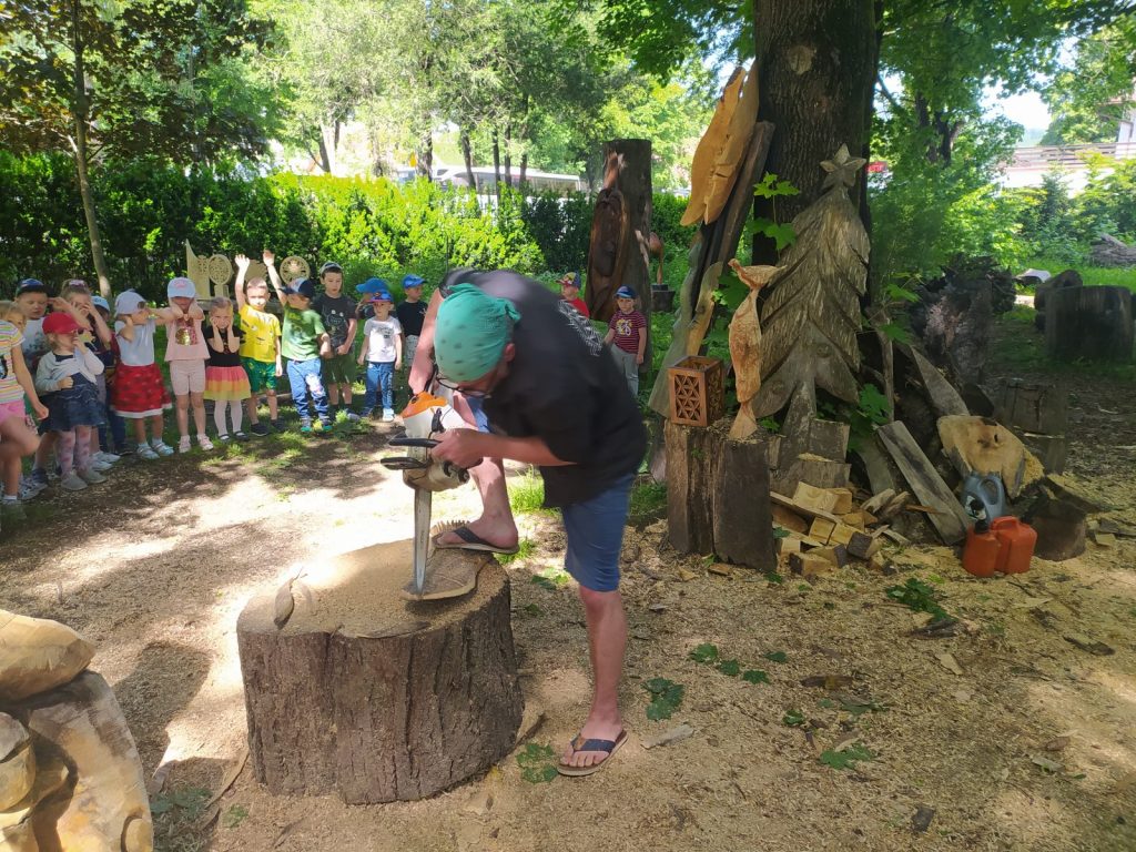 Dzieci stoją w ogrodzie.Przed dziećmi przy pniu z drzewa stoi rzeźbiarz. Z kawałka drewna rzeźbi rybę piłą motorową. Grupa w oddaleniu ogląda jego pracę. 