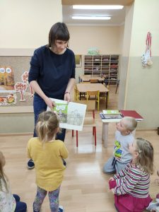 Przed dziećmi stoi ilustratorka książek i pokazuje wykonane przez nią ilustracje w książkach.