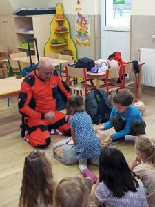 Dzieci siedzą na podłodze pezzed ratownikiem i ucza się akcji reanimacji serca