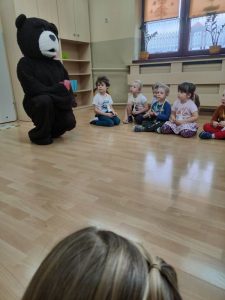 Dzieci siedzą na podłodze obok animatora w stroju misia.