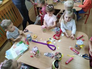 Dzieci siedzą przy stolikach i ozdabiają nakrętkę od słoika serwetką.
