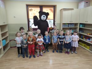 Dzieci stoją w półkolu w dwóch rzędach z tyłu stoi animator w stroju misia.