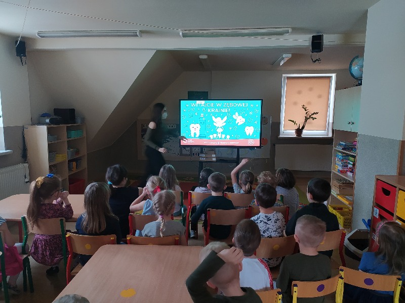 Dzieci siedzą na krzesełkach przed tablicą multimedialną .Obok tablicy Pani Stomatolog, która opowiada dzieciom o higienie jamy ustnej. Na ekranie obraz ząbków na niebieskim tle.