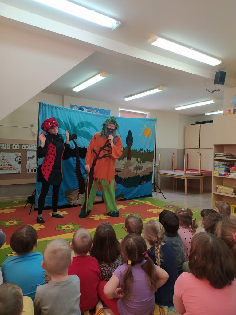 Dzieci siedzą przed sceną . Na scenie aktorzy teatru z Krakowa. Za nimi kolorowa scenografia. Aktorzy przebrani w biedronkę i skrzata 