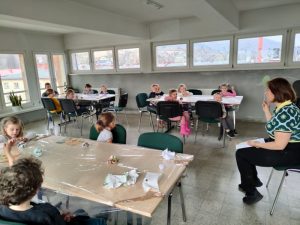 Dzieci siedzą przy stolikach 6 osobowych w bibliotece.