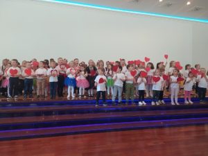 Obrazek przedstawia dużo dzieci na scenie, które trzymają w ręce czerwone serduszka