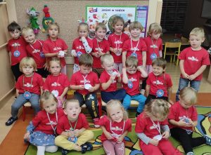 grupa maluchów ustawiona przed tablicą, dzieci w czerwonych koszulkach z medalami