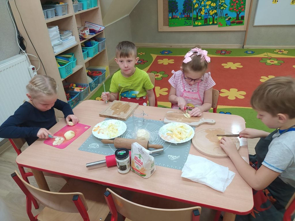 4 dzieci siedzi przy stoliku. Każde dziecko ma przed sobą deskę do krojenia nożyk, Dzieci kroją jabłka na małe kawałki i wsypuję na talerz. Każde dziecko ubrane w fartuszek