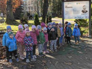 Grupa maluchów pozuje w parku do zdjęcia na tle tablicy pamiątkowej Jana Sztwiertni, na tablicy portret muzyka, krótki opis jego działalności i zasług