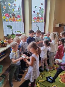 Dzieci stoją w sali , dziewczynki wręczają chłopcom prezenty.