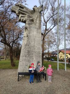 Dzieci stoją pod pomnikiem w ręku trzymają kwiaty i znicz.