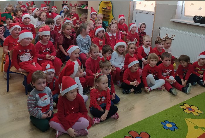 Dzieci siedzą na dywanie i na krzesełkach ubrane na czerwono. Część dzieci ma na głowie mikołajkowa czapkę a niektóre rogi renifera