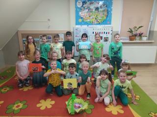 Dzieci na dywanie ubrane na zielono prezentują wykonane zadanie.Przed dziećmi koszyk ze słodyczami