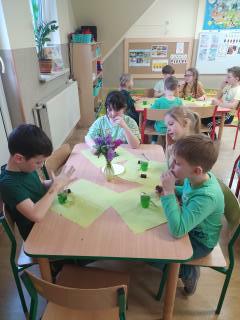 Dzieci przy stoliku jedzą zieloną galaretkę w przeźroczystych kubeczkach które znajdują się na zielonych serwetkach 