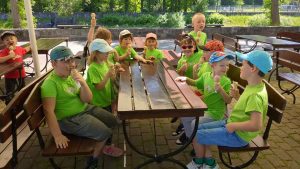 dzieci w zielonych koszulkach siedzą przy stoliku w parku i jedzą lody, niektóre mają pomalowane twarze,