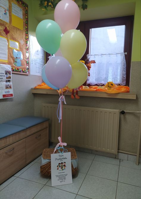 Na podłodze kosz z niespodziankami dla dzieci do którego przyczepione są baloniki oraz życzenia dla dzieci