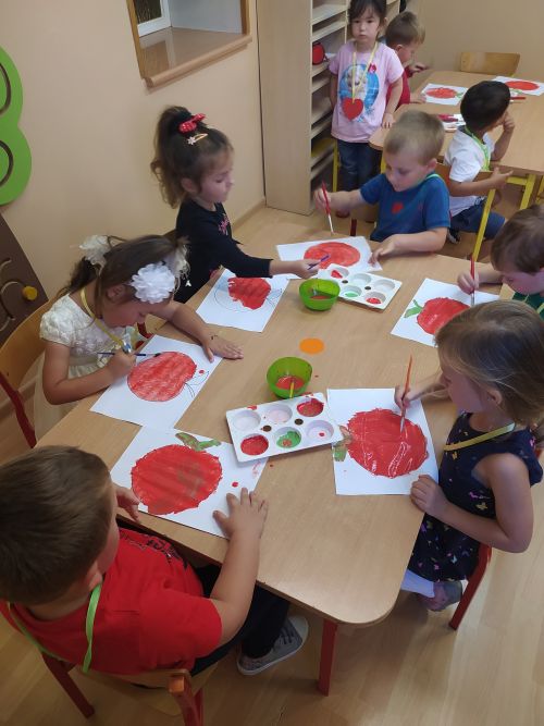 Przy stoliku siedzą dzieci i malują czerwoną farbą kontur jabłka