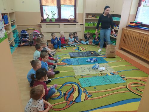 Dzieci siedzą dookoła dywanu . Na środku dywanu rozłożone 4 worki opowiadające kolorem do segregacji odpowiednich śmieci. Z prawej strony stoi prowadząca warsztaty. Dzieci segregują  "śmieci" na odpowiedni worek