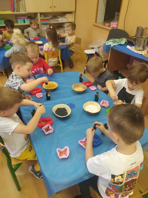 Dzieci siedzą przy stolikach na których mają plastikowe foremki. Do foremek wkładają z miseczek płatki nagietka, wiórki kokosu ,nasionka maku. Następnie do foremek wkraplają zapachowe olejki 