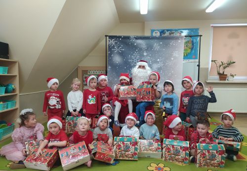 Mikołaj siedzi na krześle, obok niego z prawej i lewej strony stoją dzieci, a najmniejsze dzieci siedzą przed Mikołajem na dywanie. W większości dzieci ubrane w czerwone bluzeczki i czerwone czapeczki. Wszystkie dzieci trzymają w rękach prezent od Mikołaja