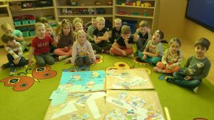 dzieci siedzą na dywanie i oglądają swoje prace plastyczne zespołowe. naszarym papierze pokolorowane , wycięte  naklejone.
