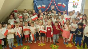 Maluchy, Średniaki, Zerówka stoją w odświętnych strojach, z flagami biało -czerwonym, w tle dekoracja patriotyczna, na materiale przypięte godło, flagi, szarfa biało -czerwona.