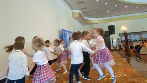 Grupa średniaków tańczy na scenie.Dzieci ustawione w kółeczkach, ubrane w dziewczynki w bluzki białe, spódniczki w kratkę różowo niebieskie