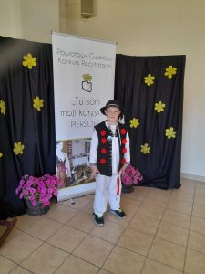 Kacper stoi w stroju regionalnym.przed tablicą z informacją o konkursie. Strój : kapelusz czarny, białe spodnie, biała koszula, bruclik-kamizelka