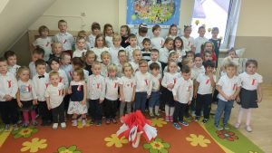 Dzieci w sali stoją uroczyście ubrane z przypiętymi patriotycznymi kotylionami patriotycznymi kotylionami, na kolorowym dywanie flagi wykonane przez dzieci i serca z papieru