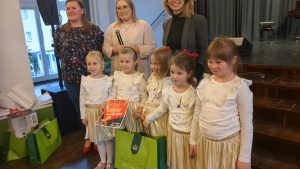 Pięć dziewczynek z naszej grupy, ubrane w białe bluzki, złote spódniczki przyjmuje nagrody w zielonej torbie idyplom, w tle jurry 