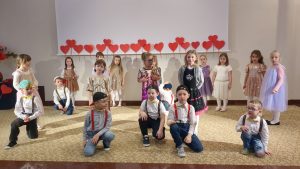 Dzieci z grupy ,,zerówki" stoją na scenie, chłopcy zprzodu, klęk prosty i pokazują swoją siłę, dziewczynki stoją z tyłu, w szeregu, w kolorowych lśniących sukienkach