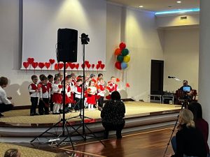 Grupa maluchów stoi na scenie, ubrana w czerwone kamizelki - chłopcy, czerwone spódniczki, białe koszulki z niebieskiej krawatem, w rękach mają serduszka