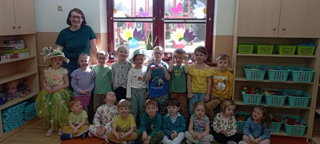 Grupa maluchów pozuje do zdjęcia, dzieci z przodu siedzą w siadzie skrzyznymy, z tyłu grupa stoi jeden obok drugiego, w grupie chłopcy i dziewczynki, dzieci ubrane w kolorze zielonym, żółtym . Jedna dziewczynka w kapeluszu, długiej sukience, stoi przed Panią w okularach i zielonej bluzce