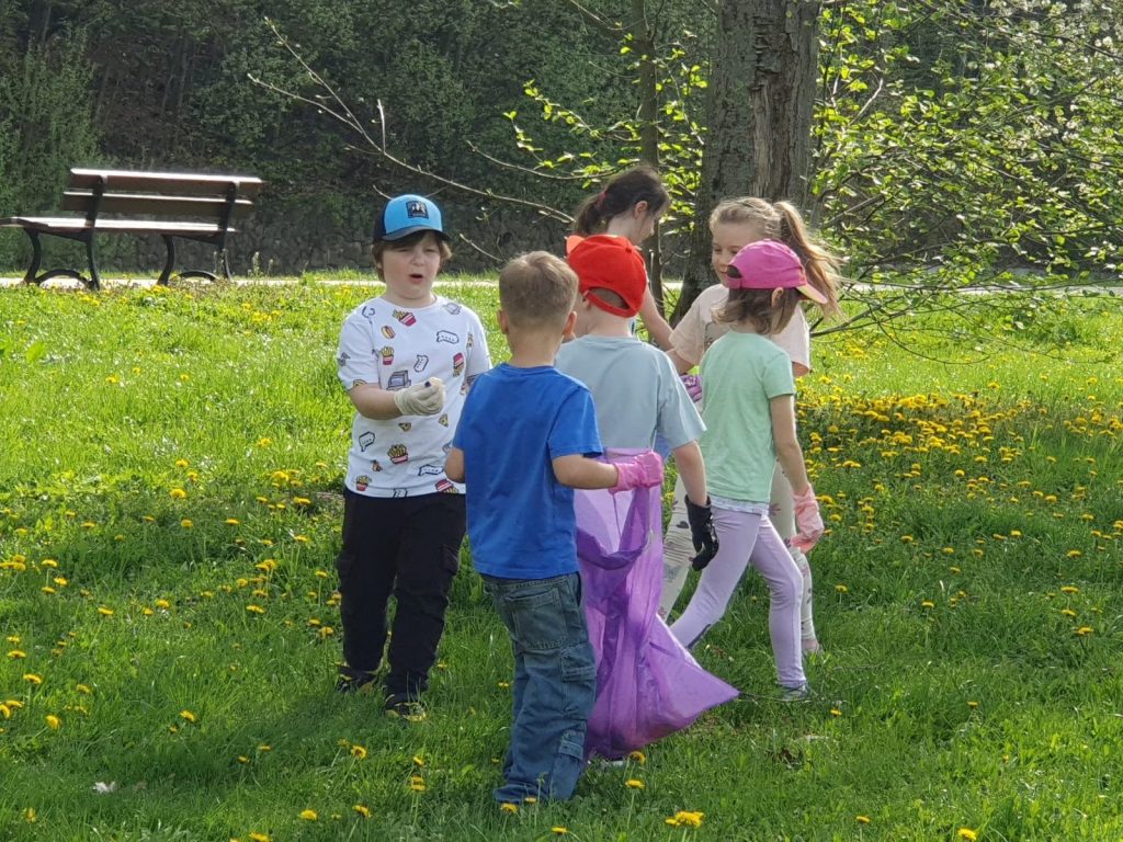 Dzieci stoją na łące, trzymają fioletowy worek na śmieci, rozglądają się dookoła.