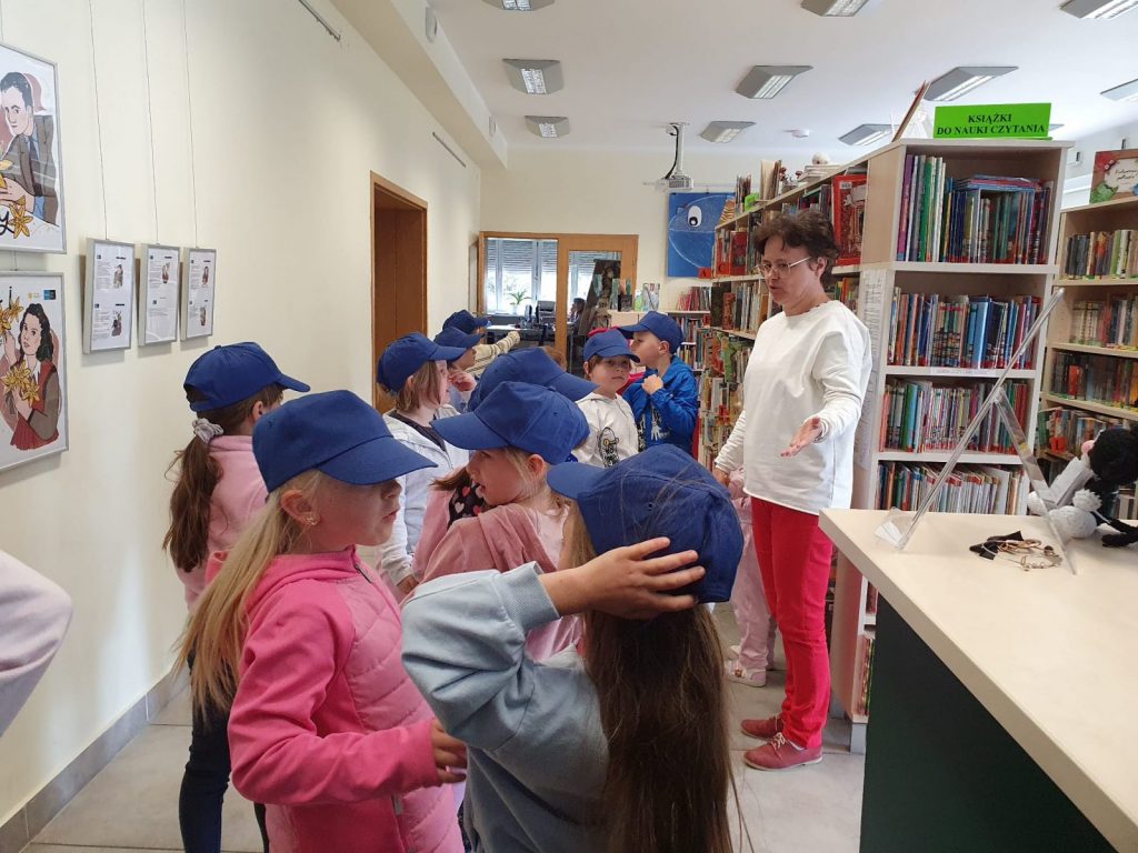 Dzieci w niebieskich czapkach stoją w bibliotece i słuchają ciekawostek o książkach, opowiada bibliotekarka 