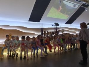 Dzieci siedzą na ławeczce w Centrum Edukacji Ekologicznej, sala jest przyciemniona, przewodnik opowiada o zasadach ekologi, 