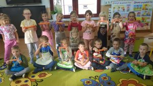 Dzieci z grupy O stoją ustawione: w rzędzie, a drugi rząd siedzie na podłodze, dzieci trzymają marchewki, a ubrane mają fartuchy