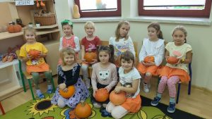 Dziewczynki z grupy strażaków w pomarańczowych ubraniach pozują siedzą na krzesłach trzymają dynie