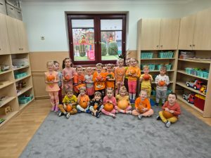 Grupa maluchów pozuje do zdjęcia, ubrani na pomarańczowo, trzymają dynie,