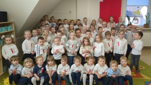 Przedszkolaki ustawione do zaśpiewania hymnu, ubrani w białe bluzki. ciemne lub czerwone spodnie i spódniczki, w tle flaga i godło 