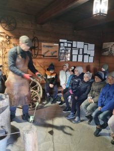 Spotkanie z kowalem w kuźni. Kowal prezentuje elementy metalowe. Dzieci siedzą na ławeczce 