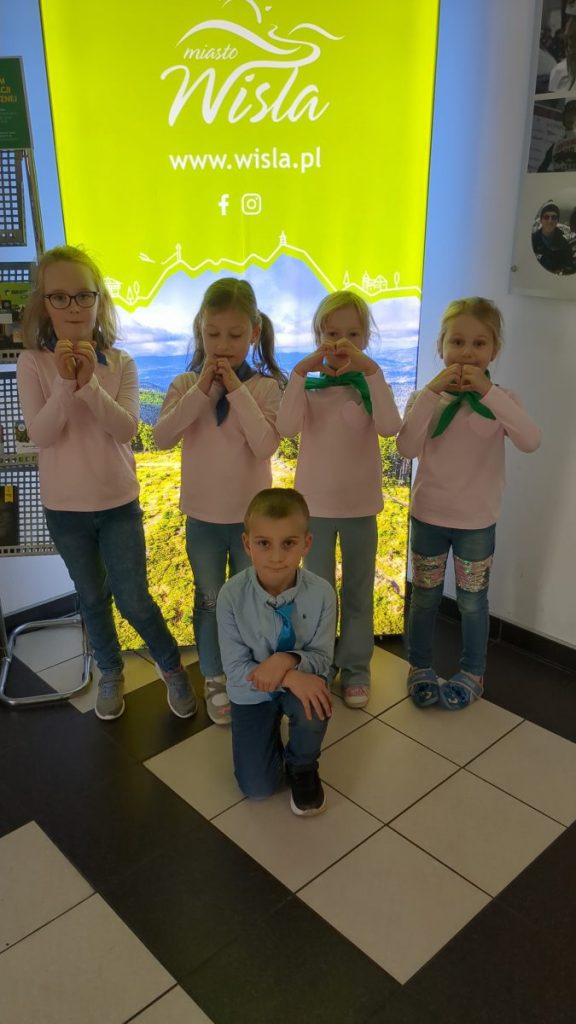 Cztery dziewczynki stoją. Pokazują serca dłońmi. Chłopak w dżinsach i niebieskiej koszuli klęczy przed dziewczynkami 