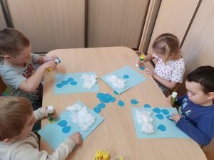 Dzieci siedzą przy stoliku i wykonują pracę plastyczną. Na niebieskich kartkach naklejają watę i kropelki deszczu z niebieskiego papieru