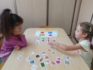 Dwie dziewczynki siedzą przy stoliku, dopasowują kształty i kolory na planszy