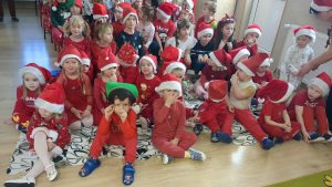 Dzieci czekają na Mikołaja. Siedzą na dywanie ubrane na czerwono, na głowach czerwone czapki.
