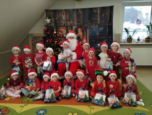 Grupa maluchów pozuje do grupowego zdjęcia z Mikołajem Mikołaj siedzi w czerwonym płaszczu, dzieci u brane na czerwono, trzymają prezenty.