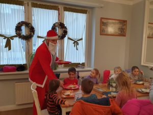 Dzieci siedzą przy okrągłym stole, ma stole dekoracje świąteczne, świeczki, bombki, gałązki. Ma czerwonych talerzach naleśniki.Dzieci jedzą, a Mikołaj stoi przed stole